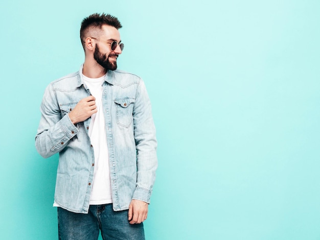 Retrato de modelo lambersexual bonito e elegante hipster homem vestido de jaqueta e jeans moda masculina posando perto da parede azul no estúdio em óculos de sol pensativo isolado