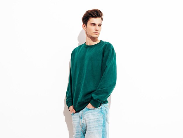 Retrato de modelo lambersexual bonito e elegante hipster homem vestido de jeans e suéter verde Moda masculina posando no estúdio perto da parede branca