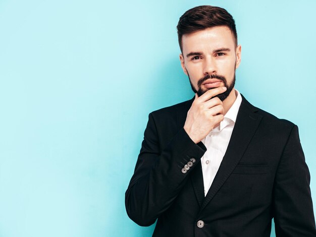 Retrato de modelo lambersexual hipster elegante confiante bonito Homem moderno sexy vestido com terno preto elegante Moda masculina posando no estúdio perto da parede azul tocando seu queixo