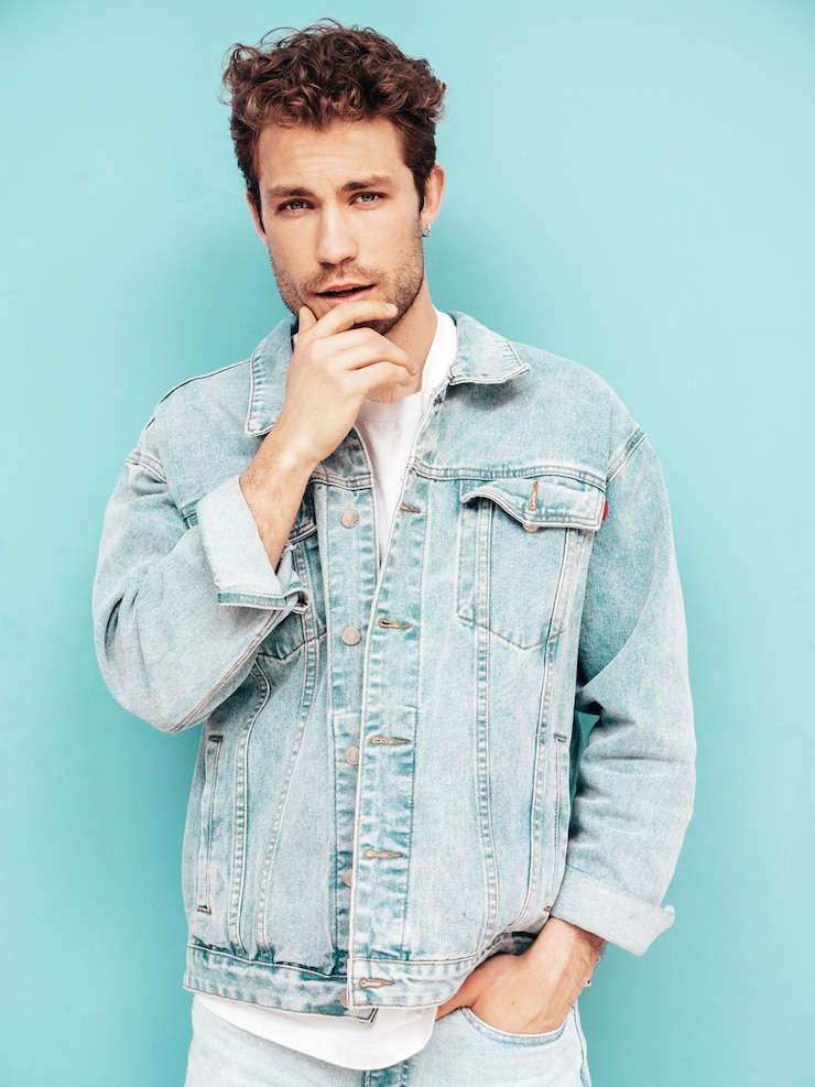 Foto grátis retrato de modelo lambersexual hipster elegante e confiante bonito com penteado encaracolado homem sexy vestido com jaqueta jeans moda masculina isolada na parede azul no estúdio tocando seu queixo