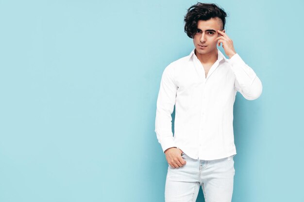 Retrato de modelo lambersexual hipster elegante e confiante bonito Homem sexy vestido com camisa branca de verão e roupas jeans Moda masculina isolada no estúdio posando perto da parede azul