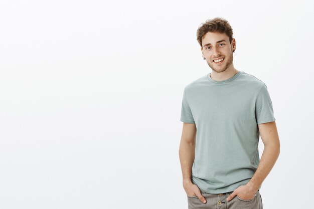 Retrato de modelo masculino atraente, despreocupado e satisfeito em roupa casual, segurando as mãos nos bolsos e sorrindo com uma expressão encantadora, inclinando a cabeça