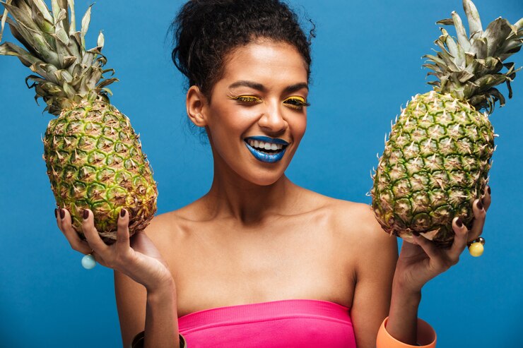 Foto grátis retrato de mulata alegre com maquiagem elegante piscando enquanto segura dois abacaxis suculentos em ambas as mãos isoladas, sobre a parede azul