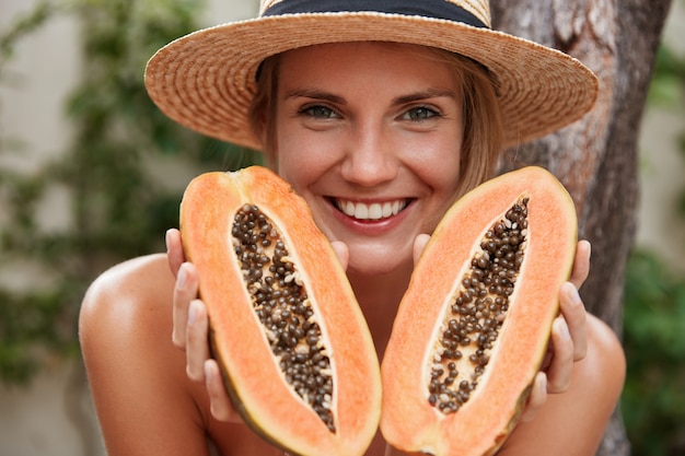 Retrato de mulher adorável feliz posa nua, usa chapéu de verão e segura mamão exótico