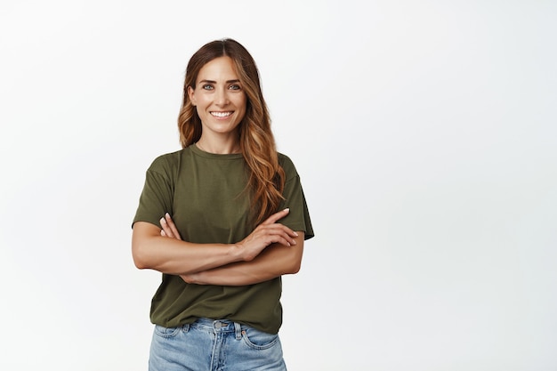 Retrato de mulher adulta confiante em t-shirt verde, de pé com os braços cruzados no peito e sorriso motivado de autoconfiança, olhando para frente, de pé contra uma parede branca.