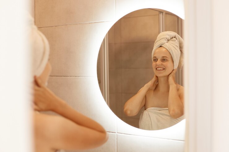 Foto grátis retrato de mulher adulta jovem fresca feliz com a pele perfeita, olhando no reflexo no espelho com um sorriso encantador, depois do banho, tocando seu pescoço, fazendo procedimentos de beleza.