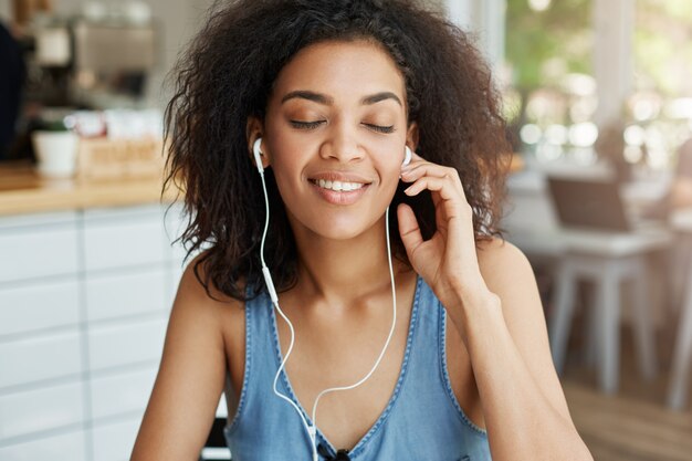 Retrato de mulher africana bonita feliz ouvindo música em fones de ouvido, sorrindo, sentado no café. Olhos fechados.