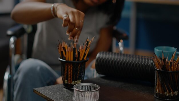 Retrato de mulher afro-americana de desenho sobre tela no espaço da oficina. Close-up de lápis de arte na mesa usada pelo jovem artista negro sentado em cadeira de rodas criando obra-prima