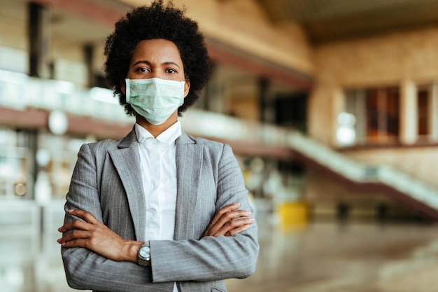 Retrato de mulher afro-americana usando máscara protetora em pé com os braços cruzados no aeroporto durante epidemia de vírus