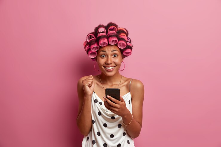Foto grátis retrato de mulher alegre de pele escura lê excelentes notícias no smartphone, levanta o punho cerrado e sorri ruidosamente, aplica rolos de cabelo para penteado. dona de casa usa redes sociais em casa
