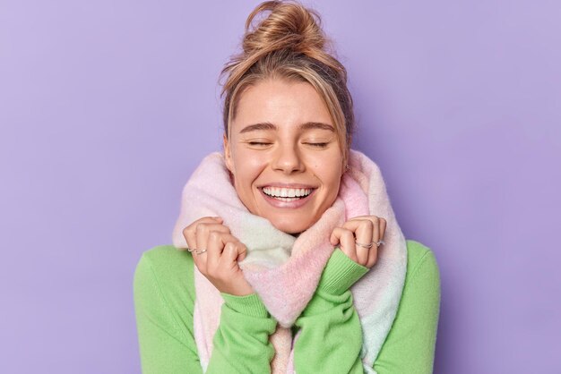 Retrato de mulher alegre gosta de suavidade de sorrisos de cachecol quente amplamente mantém os olhos fechados tem cabelo penteado se veste para o inverno isolado sobre fundo roxo. Emoções sinceras felizes