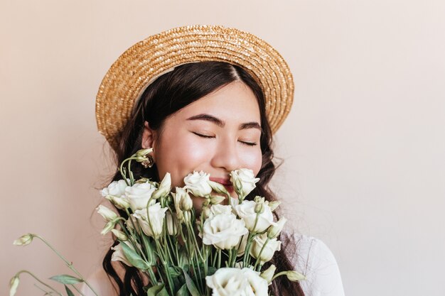 Retrato de mulher asiática com chapéu de palha cheirando flores com os olhos fechados. Foto de estúdio da bela mulher japonesa segurando buquê de eustomas brancos.