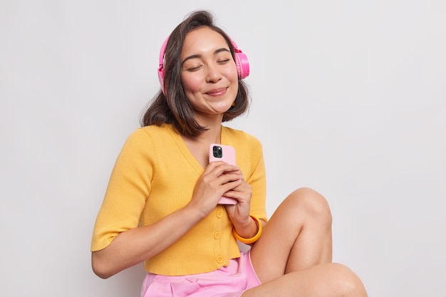 Retrato de mulher asiática satisfeita com cabelo escuro curtindo música favorita som perfeito em fones de ouvido sem fio mantém celular moderno mantém os olhos fechados usa roupas casuais isoladas sobre a parede branca