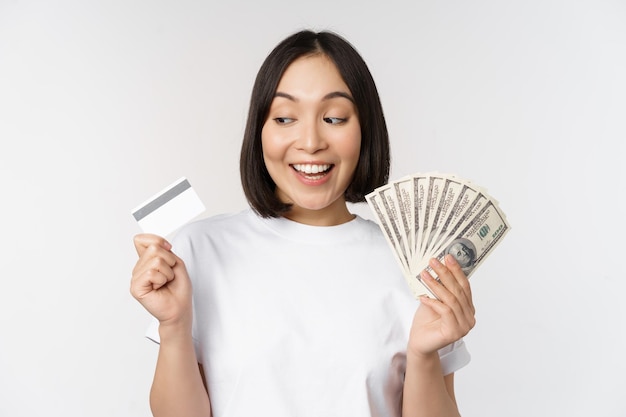 Retrato de mulher asiática sorrindo segurando cartão de crédito e dólares em dinheiro em pé na camiseta sobre fundo branco