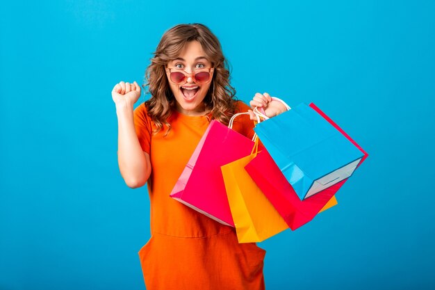 Retrato de mulher atraente animada, sorridente e elegante, shopaholic em um vestido laranja da moda, segurando sacolas de compras no fundo azul do estúdio isolado