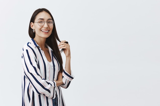 Retrato de mulher atraente, despreocupada e feliz, de óculos e blusa listrada, brincando com uma mecha de cabelo e sorrindo com confiança