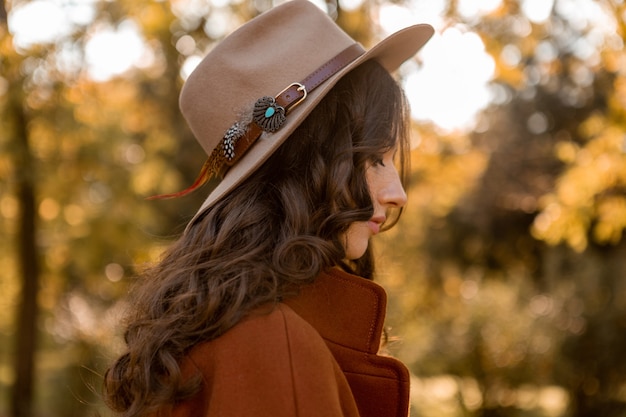 Retrato de mulher atraente e elegante com cabelo longo encaracolado caminhando no parque vestida com casaco marrom quente outono na moda, estilo de rua usando chapéu