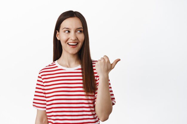 Retrato de mulher bonita apontando o dedo para a direita, sorrindo e olhando para o logotipo, contando sobre um grande evento de venda, descontos na loja, em pé contra uma parede branca