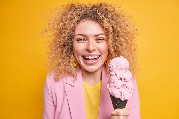 Retrato de mulher bonita com cabelo encaracolado tem sorvete de cone tem passeios de dia de folga com amigos no parque durante o verão isolado sobre a parede amarela. modelo feminina comendo sobremesa gostosa