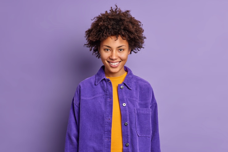 Foto grátis retrato de mulher bonita de cabelo encaracolado sorri alegremente usa elegante jaqueta roxa em um tom com fundo se sente encantado poses contentes interior fala com um colega de trabalho. conceito de emoções positivas