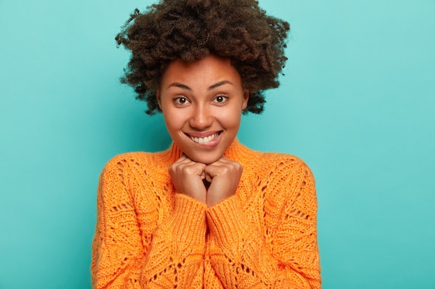 Retrato de mulher bonita e cacheada morde os lábios, mantém as mãos embaixo do queixo, olha com alegria e adoração, morde os lábios, sorrindo com os dentes, usa suéter de tricô, recebe notícias agradáveis, estande interior