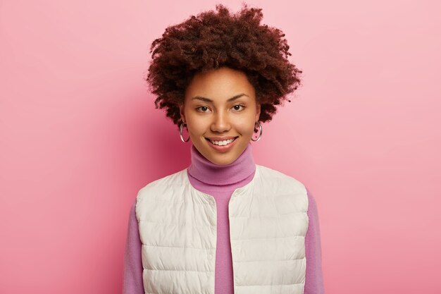 Retrato de mulher bonita encaracolada tem beleza natural, sorri agradavelmente, usa brincos, colete branco, expressa alegria, posa contra um fundo rosa.