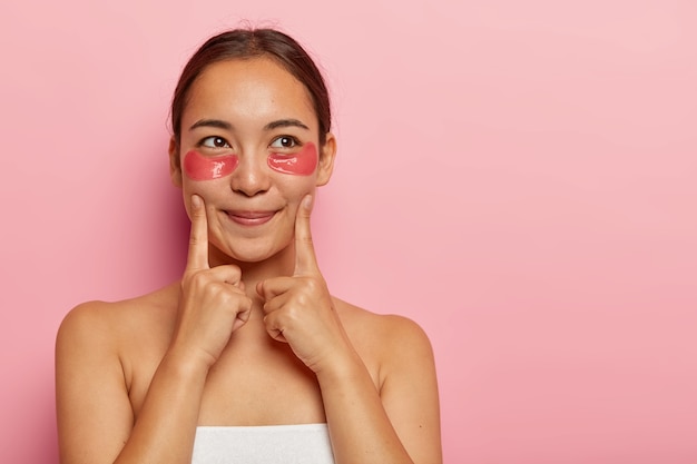 Retrato de mulher bonita tem pele fresca, aponta para as bochechas, tem manchas de hidrogel embaixo dos olhos, aplica máscara de colágeno anti-rugas, fica enrolado em toalha, olha de lado, isolado na parede rosa. Beleza