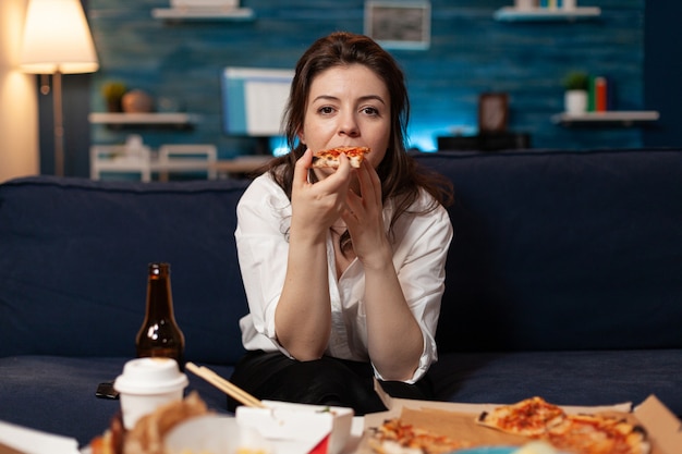 Retrato de mulher branca, sentada no sofá, comendo uma fatia de pizza, desfrutando de comida para viagem em casa ...