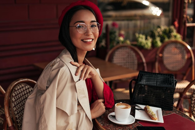 Foto grátis retrato de mulher bronzeada asiática em vestido vermelho elegante boina bege trench coat e óculos senta-se em aconchegantes sorrisos de café de rua e gosta de sobremesa e café