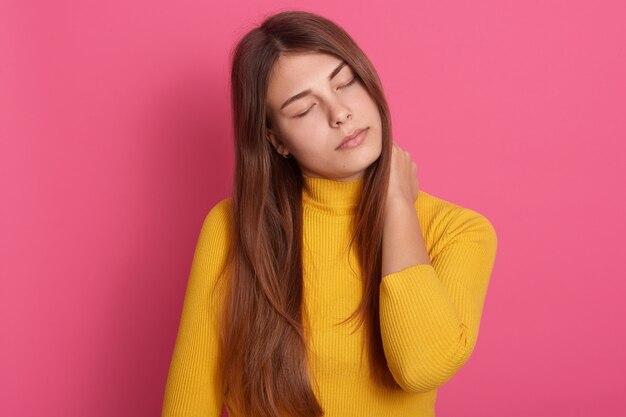 Retrato de mulher cansada e doente com cabelo comprido, vestindo uma camisa amarela casual, em pé com os olhos fechados, sentindo dores no pescoço, massageando os músculos tensos