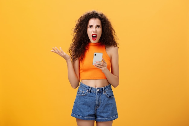 Retrato de mulher caucasiana chateada e irritada descontente com penteado encaracolado em batom vermelho e top laranja recortado segurando smartphone e gesticulando com a palma da mão irritado reclamando sobre mensagem idiota.