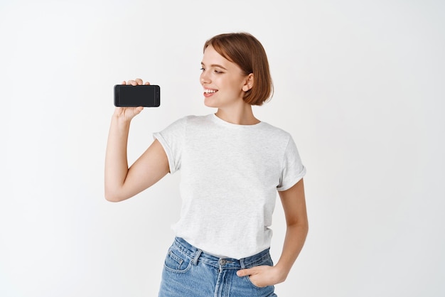 Retrato de mulher caucasiana sorridente com cabelo curto, mostrando a tela do smartphone e olhando para ele em pé contra um fundo branco