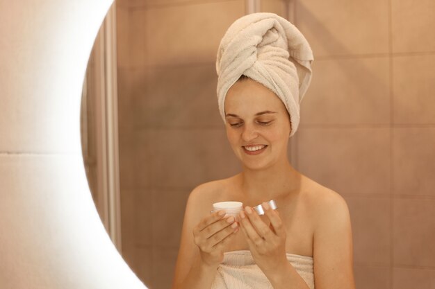 Retrato de mulher com creme nas mãos, reflexo de mulher sorridente com uma toalha na cabeça, fazendo procedimentos de beleza em casa no banheiro, cuidando da pele dela.