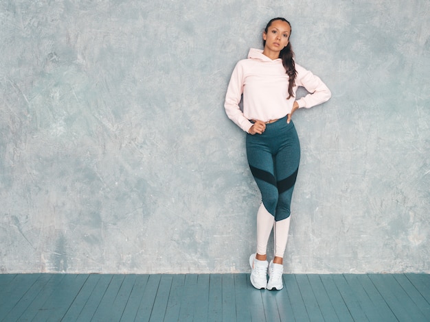 Retrato de mulher confiante fitness em roupas esportivas, olhando confiante. Feminino posando no estúdio perto da parede cinza