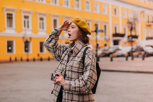 Foto grátis retrato de mulher de boina amarela com câmera retrô nas mãos garota de casaco xadrez olha para magníficos edifícios europeus com surpresa e prazer