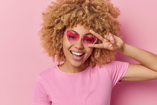 Retrato de mulher de cabelo encaracolado feliz faz gesto de paz sobre sorrisos de dente tem expressão feliz tolos ao redor usa óculos de sol de coração e camiseta casual isolada sobre fundo rosa