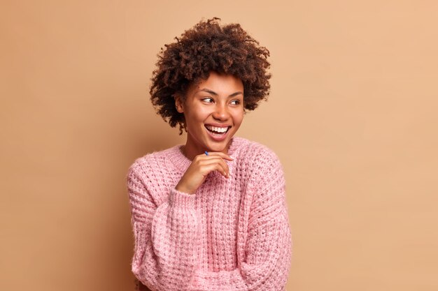 Retrato de mulher de cabelo encaracolado mantém a mão sob o queixo e desvia o olhar alegremente usando um macacão de malha e poses de expressão despreocupada contra a parede marrom