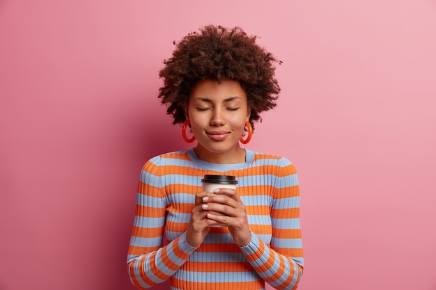 Retrato de mulher de cabelos cacheados adora café, segura copo descartável de bebida, fecha os olhos de prazer e satisfação, usa blusa listrada, fica encostada na parede rosa, tem sonhos agradáveis