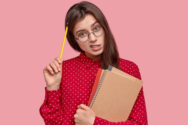 Retrato de mulher de camisa vermelha e óculos redondos segurando blocos de notas