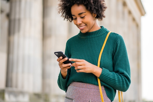 Retrato de mulher de negócios afro usando seu telefone celular em pé ao ar livre na rua