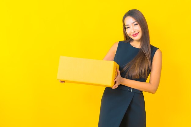 Retrato de mulher de negócios asiática jovem bonita com caixa marrom pronta para envio em fundo amarelo.