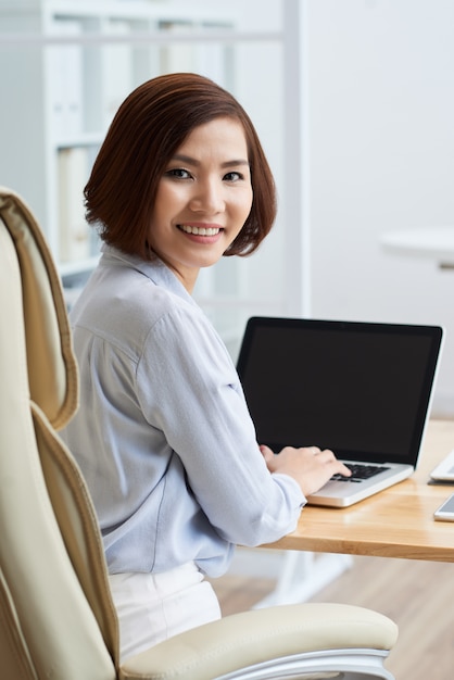 Retrato de mulher de negócios, voltando-se para olhar para a câmera com as mãos digitando no teclado na mesa do escritório