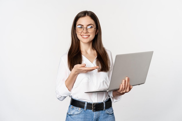 Retrato de mulher de óculos segurando laptop apontando para tela mostrando seu trabalho no computador em pé...