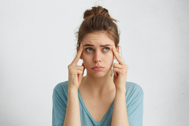 Retrato de mulher de olhos azuis caucasiana cansada com cabelo loiro, segurando os dedos nas têmporas, olhando para cima tendo olhar frustrado, tentando se lembrar de algo importante. Mulher cansada pensando em descansar