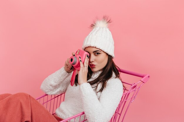 Retrato de mulher de olhos castanhos no chapéu branco e suéter tirando foto na instax enquanto está sentado no carrinho de supermercado na parede rosa.