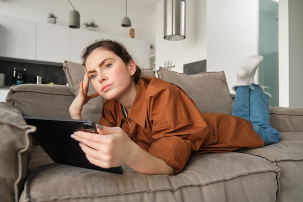 Retrato de mulher deitada no sofá em casa segurando um tablet digital e olhando pensativo franzindo a testa