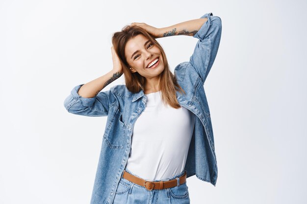 Retrato de mulher despreocupada tocando seu cabelo e inclinando a cabeça feliz, sorrindo, se divertindo otimista, em pé no branco