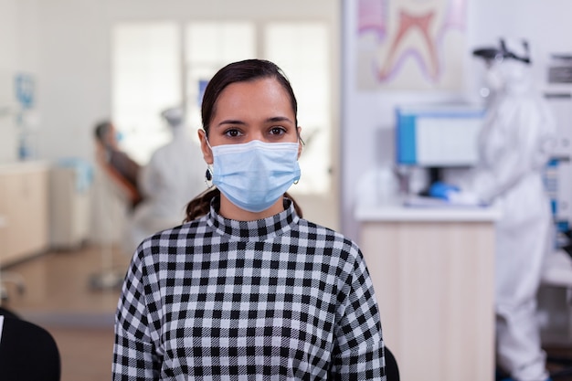 Retrato de mulher em consultório odontológico, olhando para a câmera usando máscara facial, sentado na cadeira na clínica de sala de espera enquanto o médico trabalha