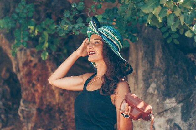 Retrato de mulher em pé e segurando seus chapéu e camerand em camisa preta na beira-mar durante o dia.