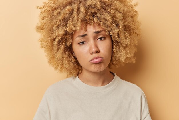 Retrato de mulher encaracolada triste frustrada parece infelizmente sente muito pelo que aconteceu bolsas lábios vestidos com camiseta casual isolada sobre fundo marrom Conceito de pessoas e emoções negativas